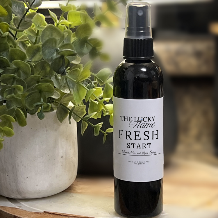 Fresh Start room, linen, and car spray with soft baby powder, rose, and sweet orange notes in a sleek 4 oz black bottle styled with white towels and soft pink florals.