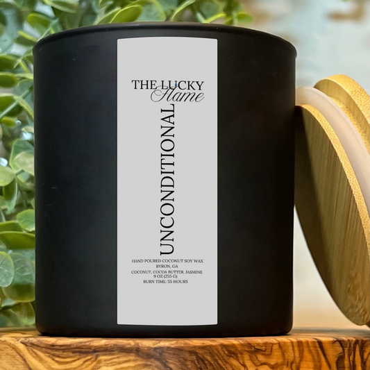 Unconditional candle in a sleek 9 oz black jar with notes of coconut, cocoa butter, and jasmine, styled with soft textures and warm, calming lighting.